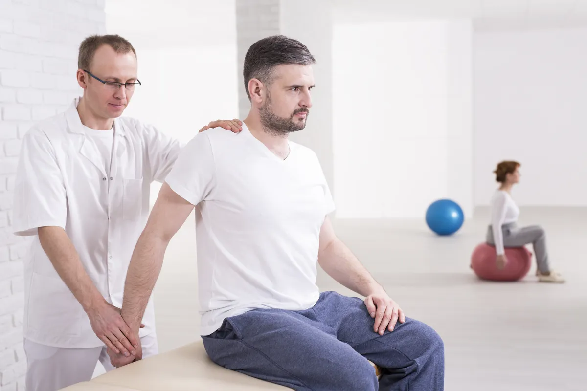 Man during rehabilitation and specialist helping him during exercises, sitting in light interior