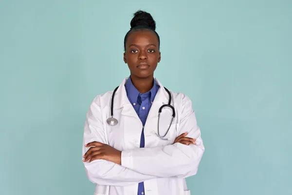 Confident african female doctor wear white lab coat, stethoscope look at camera stand on mint green studio background. Serious proud black woman professional therapist physician arms crossed portrait.