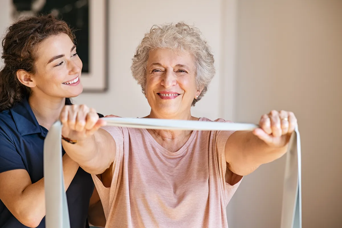 Personal trainer assisting senior woman with resistance band. Rehabilitation physiotherapy worker helping old patient at nursing home. Old woman with stretch band being coached by physiotherapist.